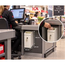 Vit sedelbox med nyckellås monterad vid kassadisk i butik. En hand stoppar ner en sedel i boxens övre öppning.