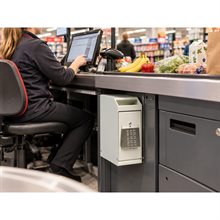 Ljusgrå sedelbox med numerisk kodpanel monterad på en grå kassadisk i en butiksmiljö, med en person vid kassan.